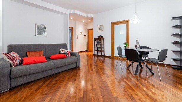 A modern living room with a grey sofa, red cushions, a dining table with chairs, and a wooden floor. - GuestHost - Roomy and tastefully furnished apartment, ideal for families as it accommodates up to 6 people having 2 bedrooms, 2 bathrooms, a balcony and an equipped terrace. The flat is located in a residential context with concierge and internal garden, (Bari)