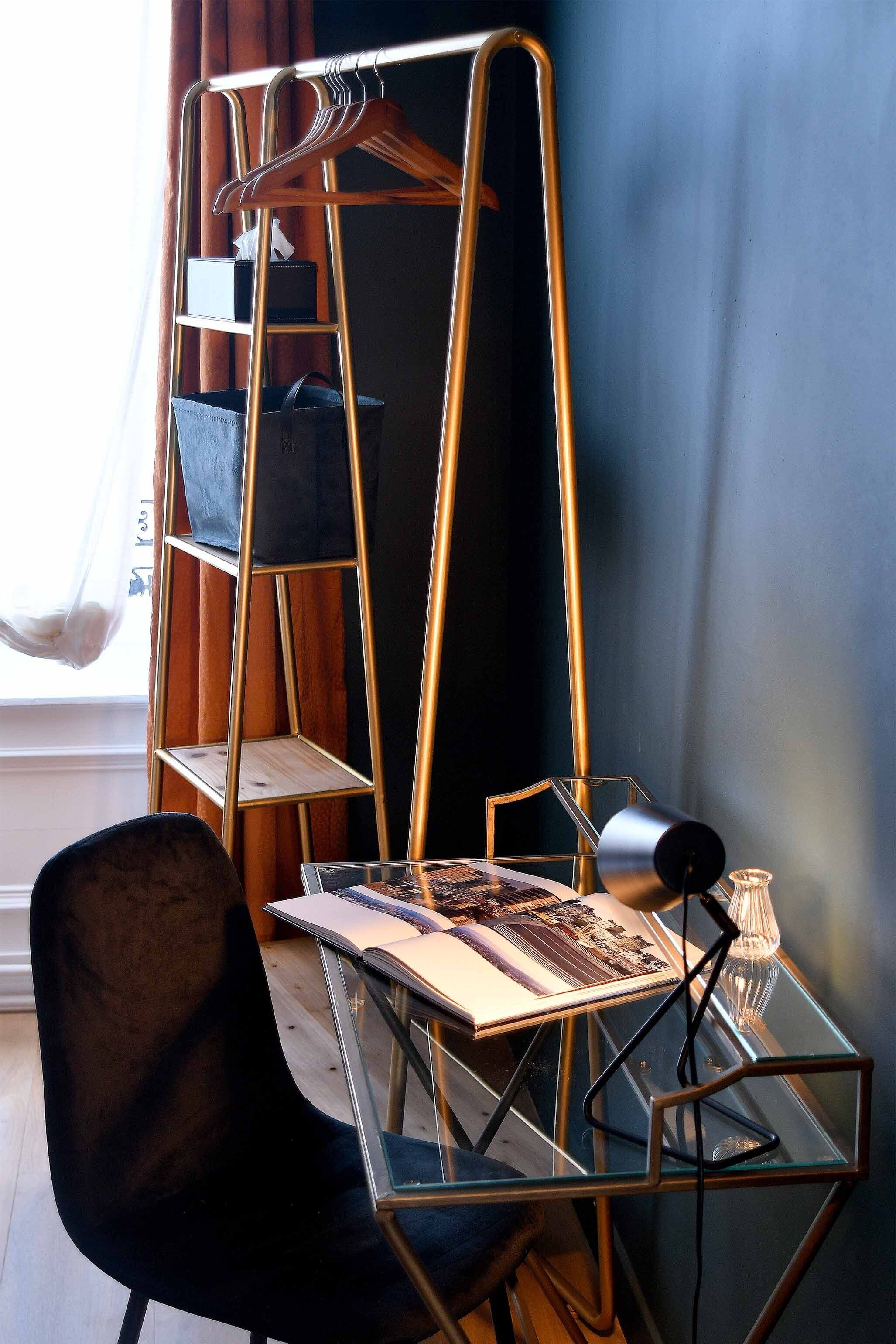 A modern room with a glass table, a black chair, a gold ladder shelf, and a window with curtains.