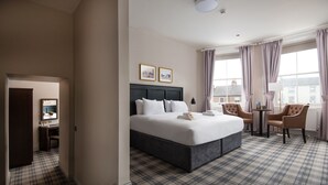 A hotel room with a large bed, a desk, and a chair. There are two framed pictures on the wall, a window with curtains, and a plaid carpet. - The Northallerton Inn - The Inn Collection Group (Northallerton)