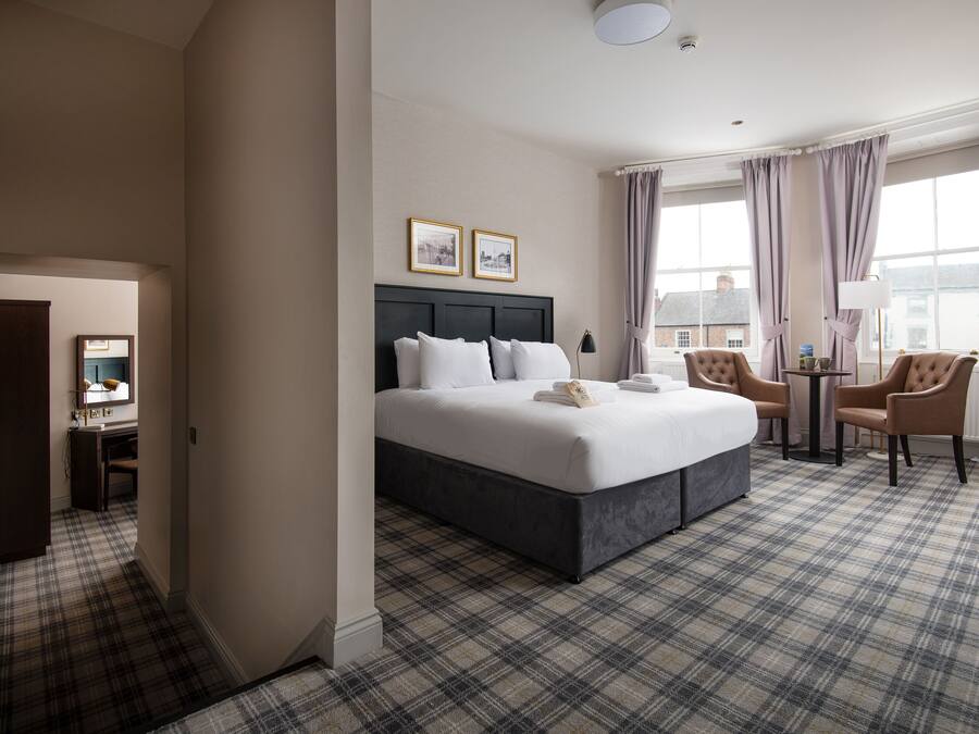 A hotel room with a large bed, a desk, and a chair. There are two framed pictures on the wall, a window with curtains, and a plaid carpet.
