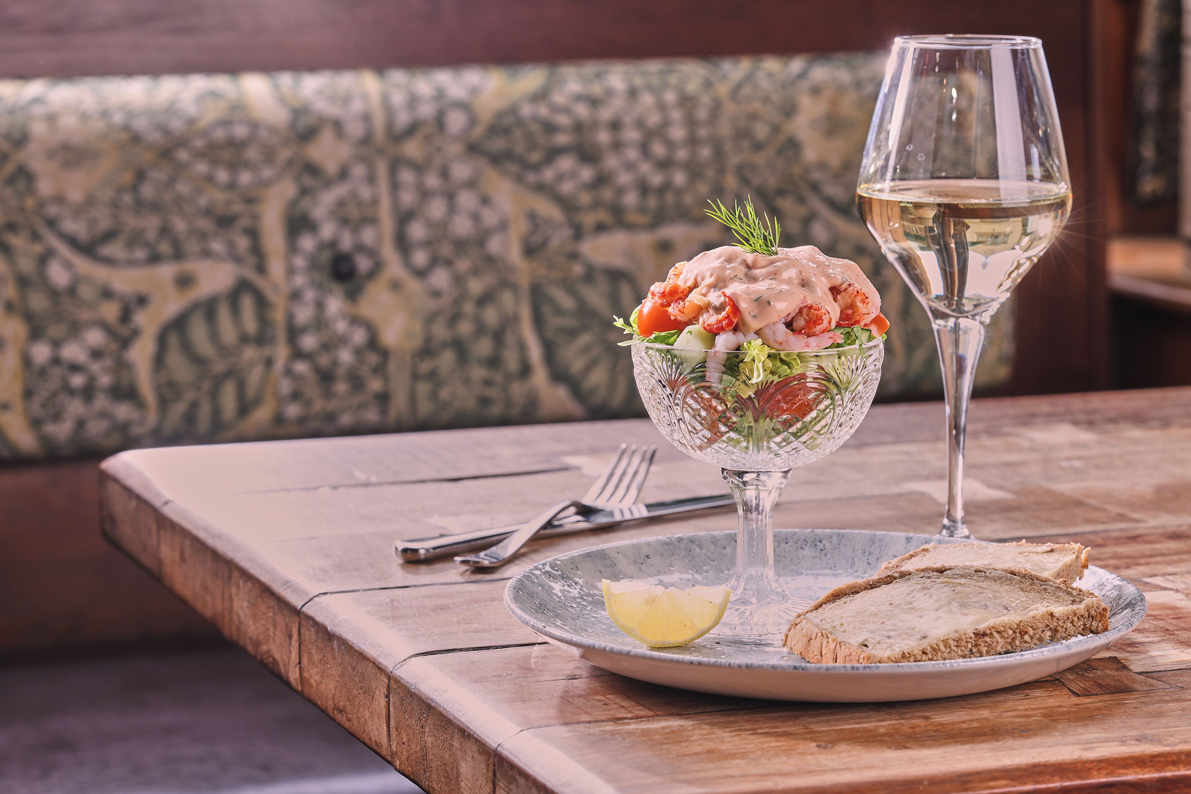 A dining table with a glass salad bowl, a glass of white wine, and a slice of bread on a plate.
