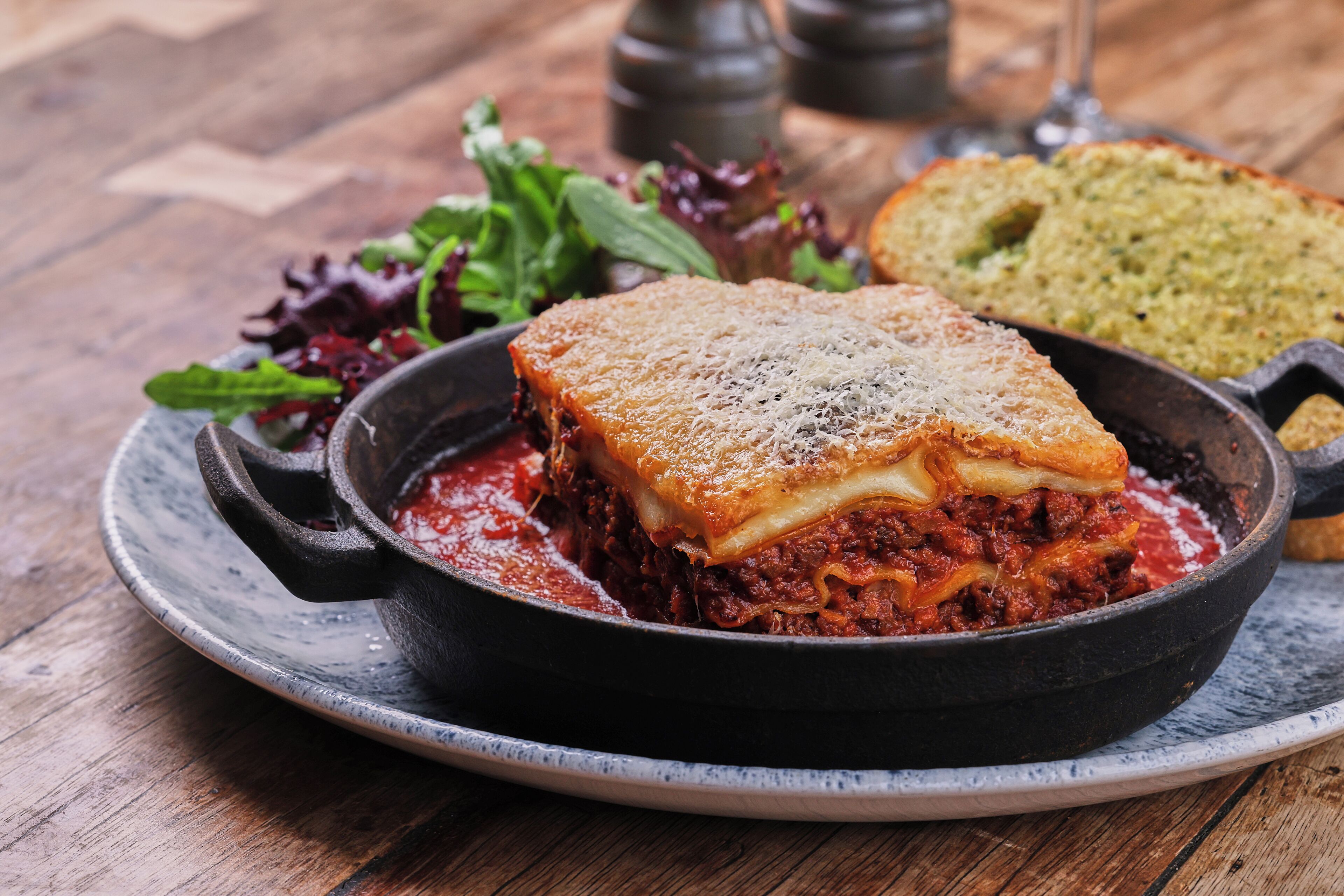 A dish of lasagna in a cast iron skillet on a plate, accompanied by a side salad and a piece of bread.