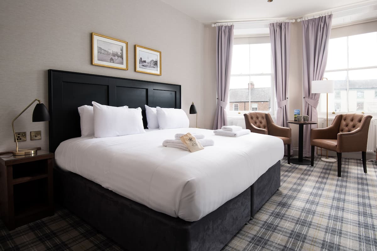 A hotel room with a large bed, two armchairs, a small table, and a window with curtains.