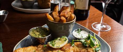 A plate of fish and chips with sides of mushy peas and tartar sauce, a glass of red wine, and a bottle of wine on a wooden table.