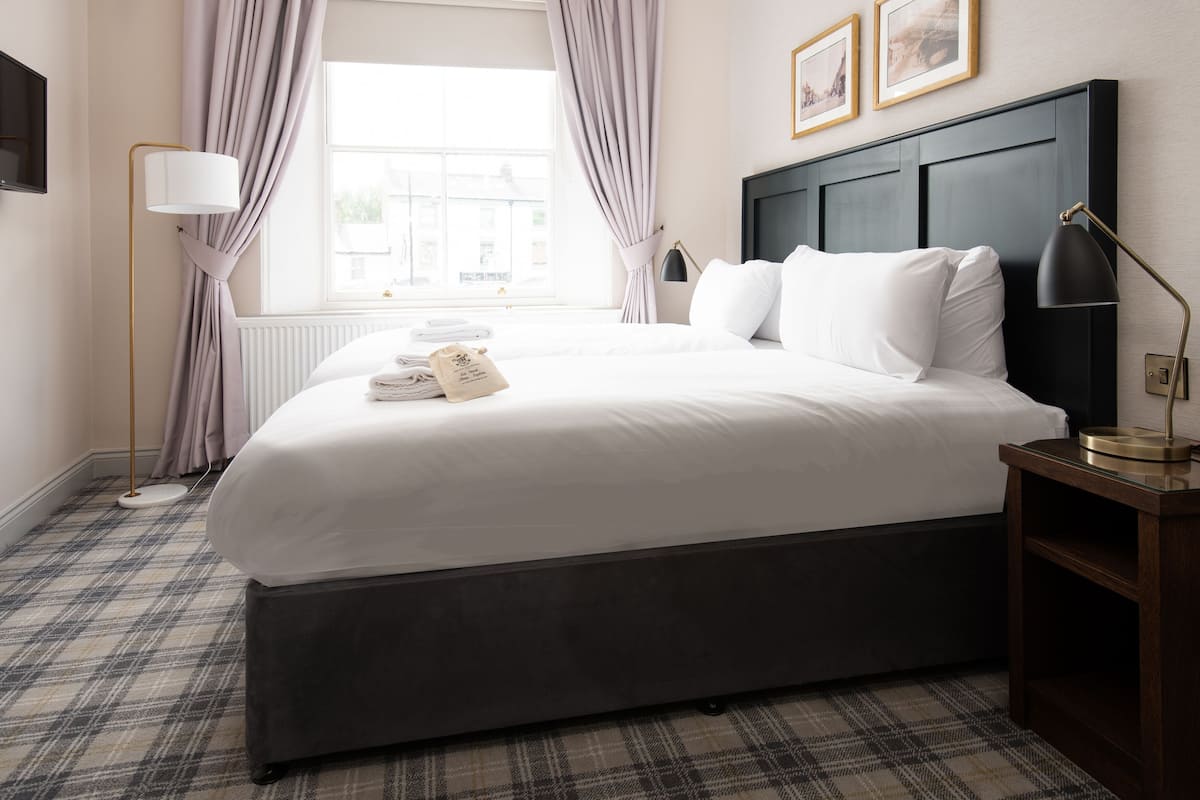A hotel room with a large bed, a headboard, a bedside table with a lamp, a floor lamp, and a window with curtains.