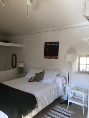 A bedroom with a bed, pillows, a bedside table with a lamp, a small stool, a window, and a wall-mounted artwork.