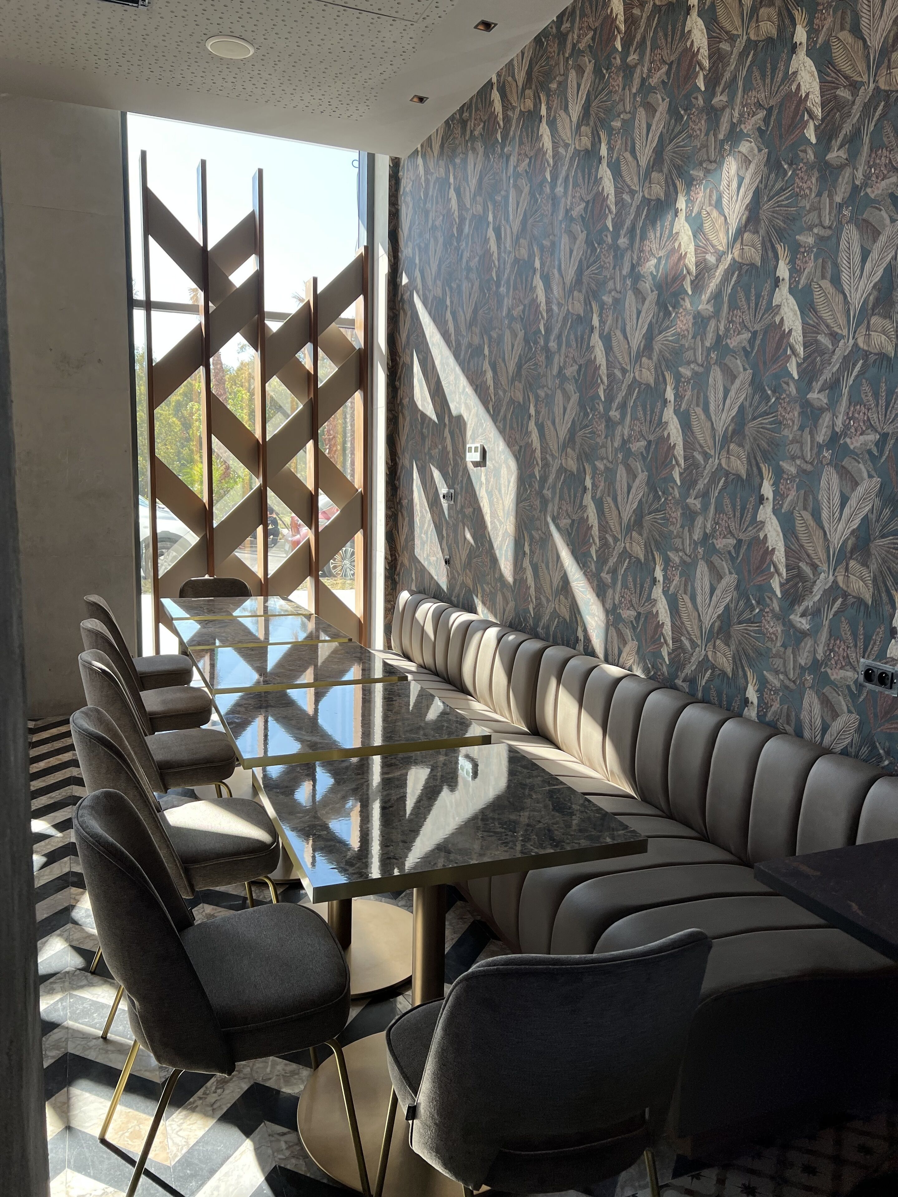 A modern dining area with a long table and cushioned seating, a patterned wall, and a view through a lattice window.