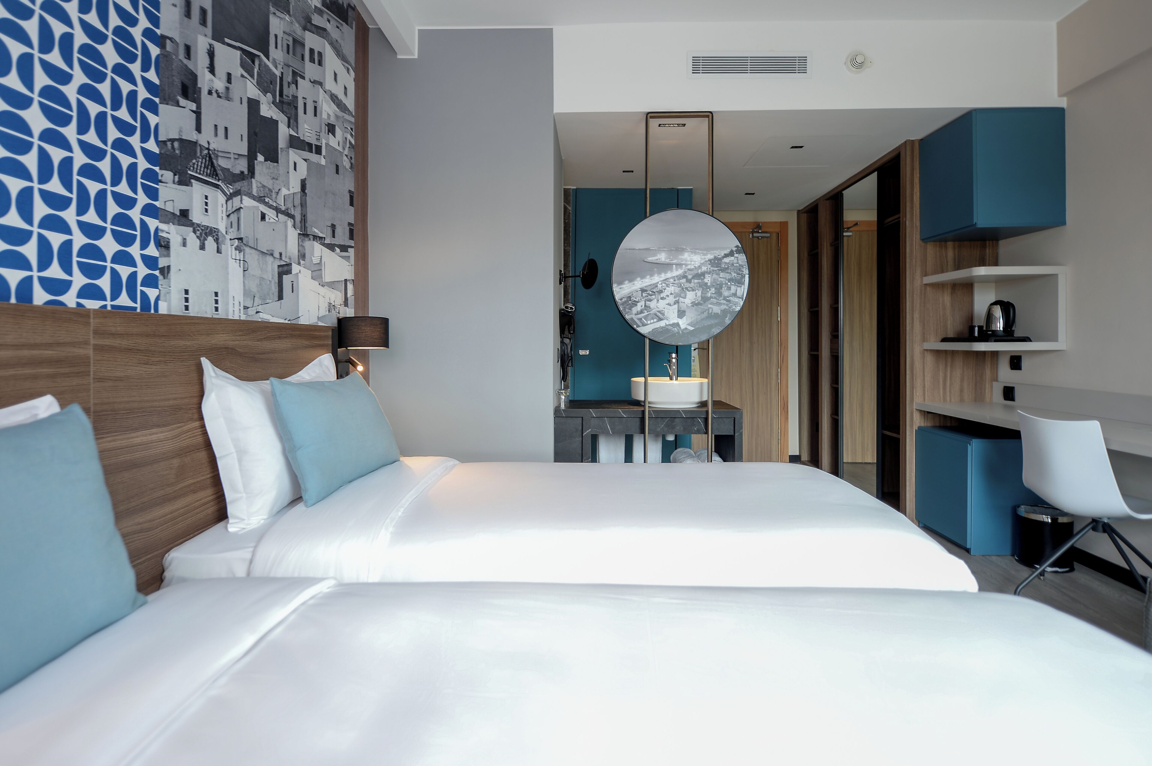 A modern hotel room with a large bed, a desk with a mirror, and a view of a cityscape through a large window.