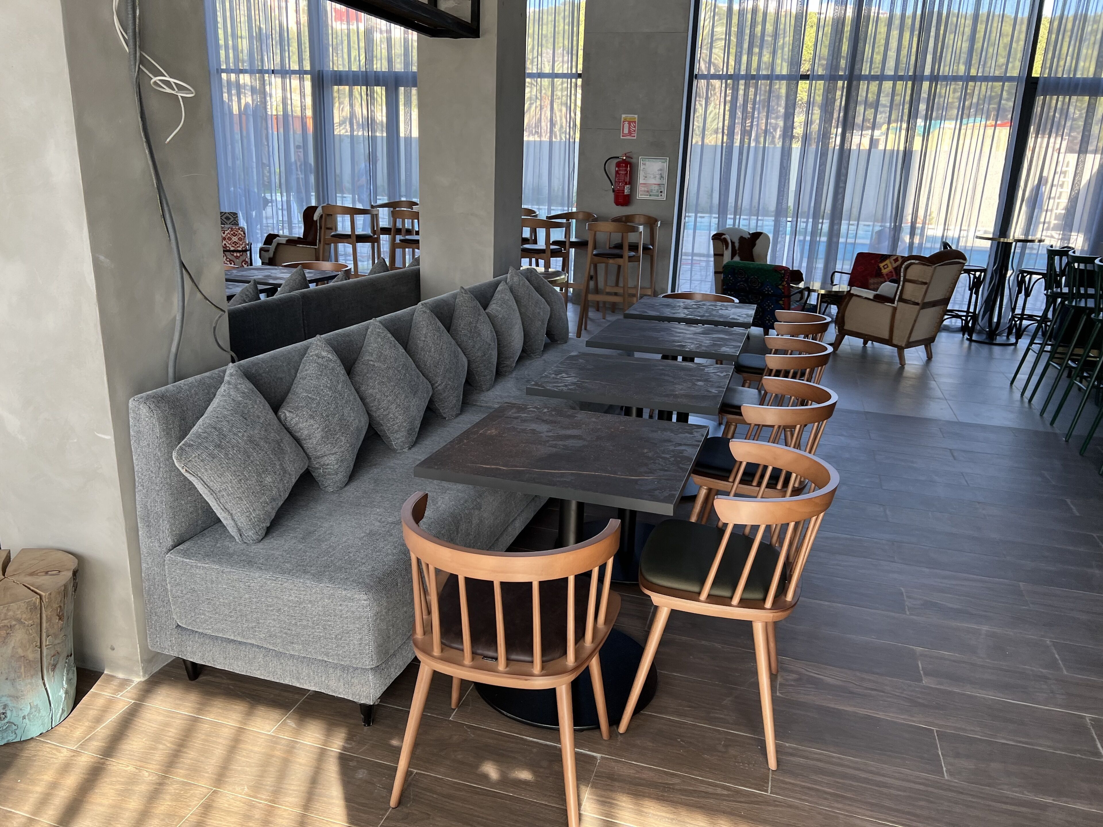A modern dining area with a long grey sofa and wooden chairs, large windows with sheer curtains, and a fire extinguisher on the wall.