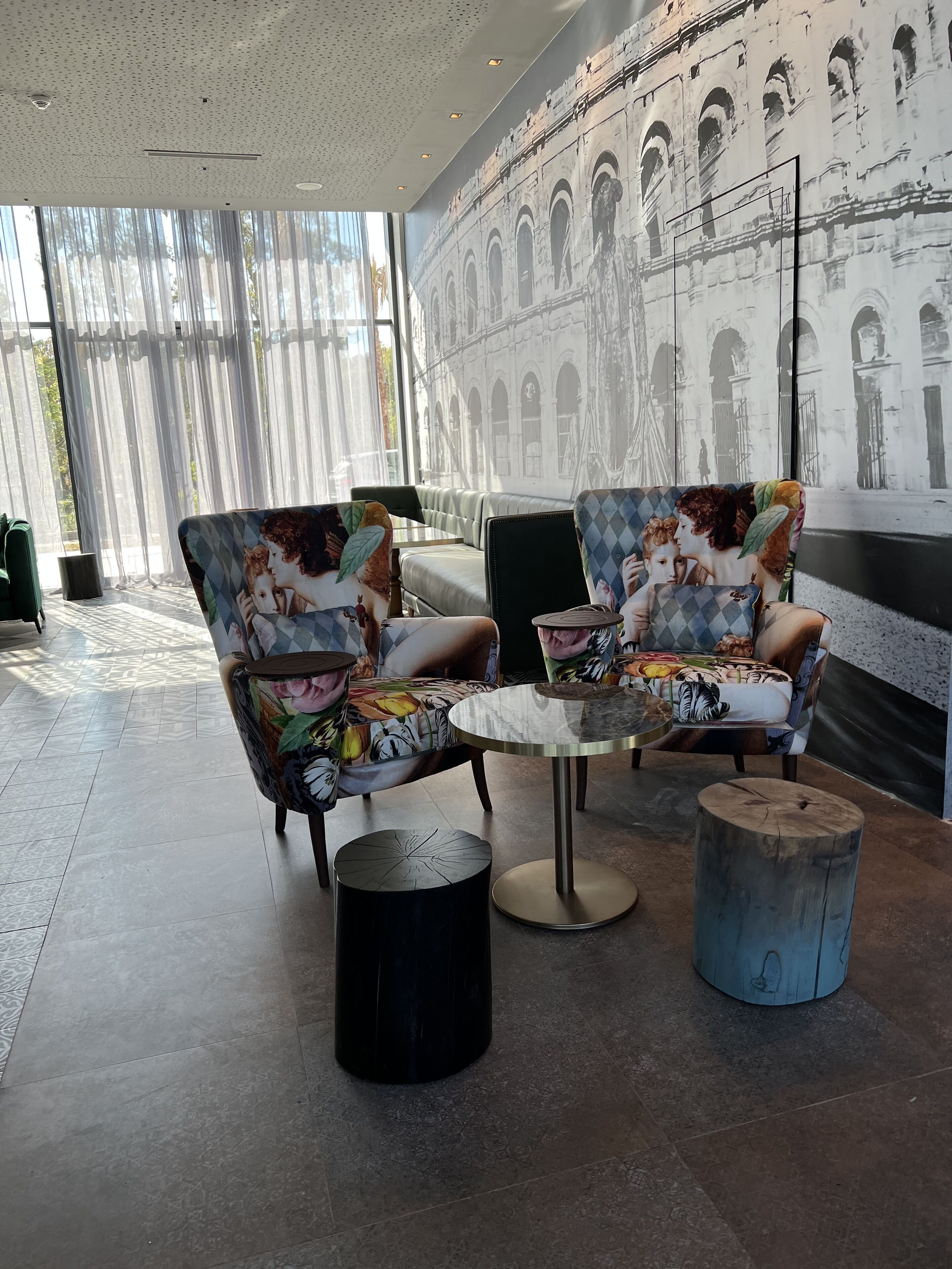 A modern lounge area with patterned armchairs, a glass-top coffee table, and a mural of an architectural structure.