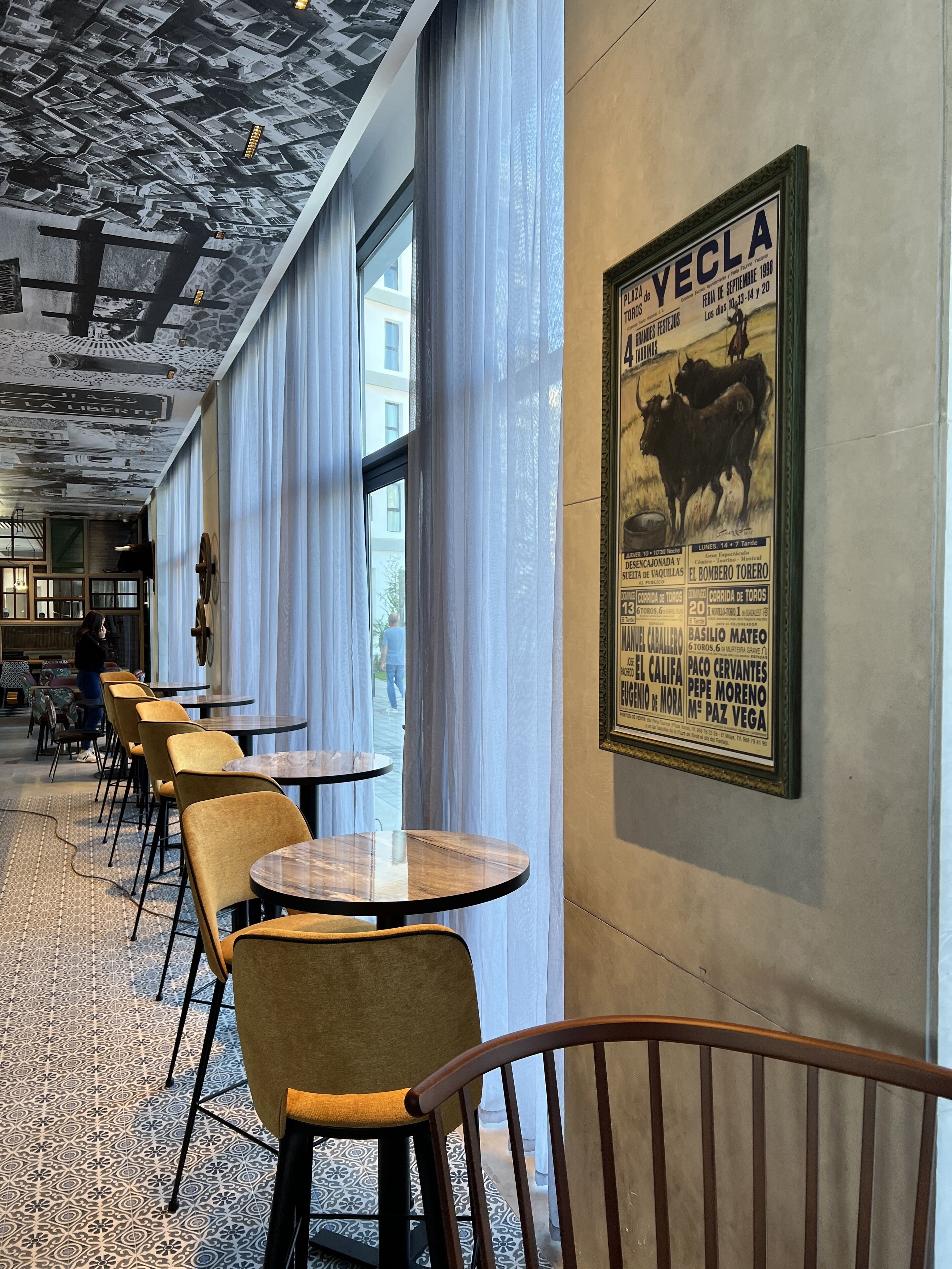 Un restaurant avec des tables et des chaises rondes, une fresque murale et une grande affiche faisant la promotion des événements locaux.