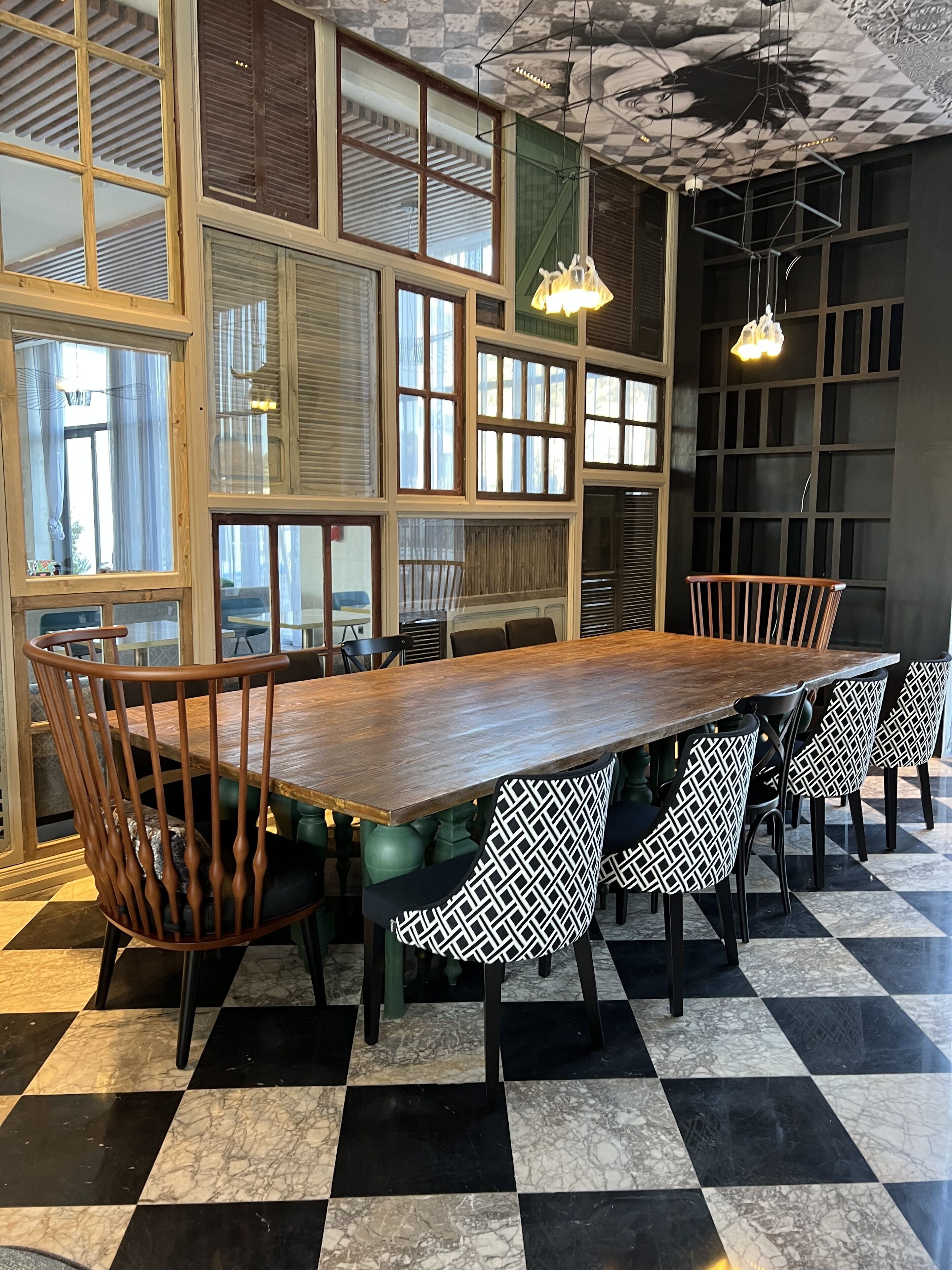 A dining area with a large wooden table surrounded by chairs, large windows with blinds, and a checkered floor.