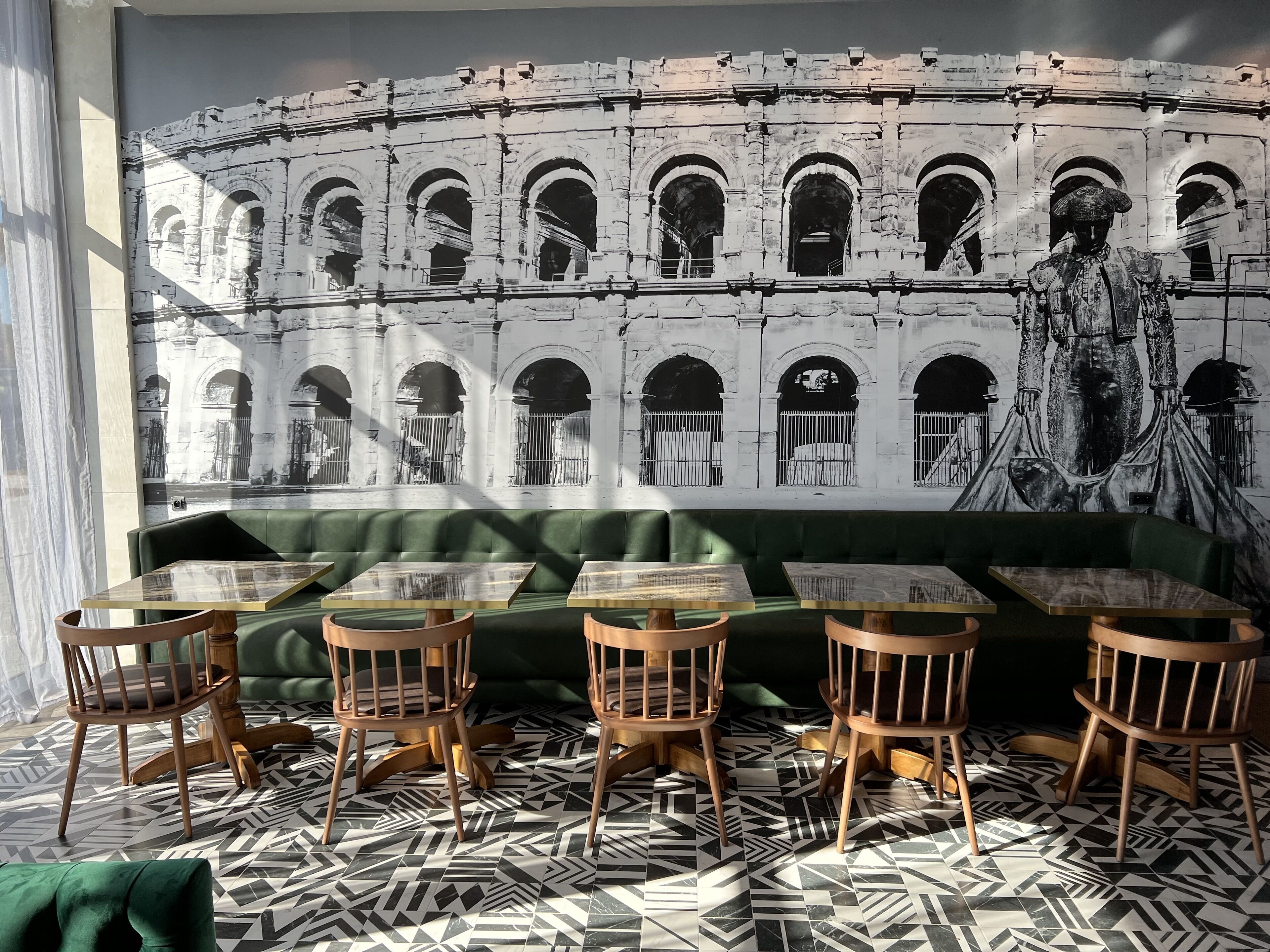 Un restaurant décoré d’une fresque représentant un ancien amphithéâtre, avec des sièges rembourrés de couleur verte et des tables et chaises en bois.