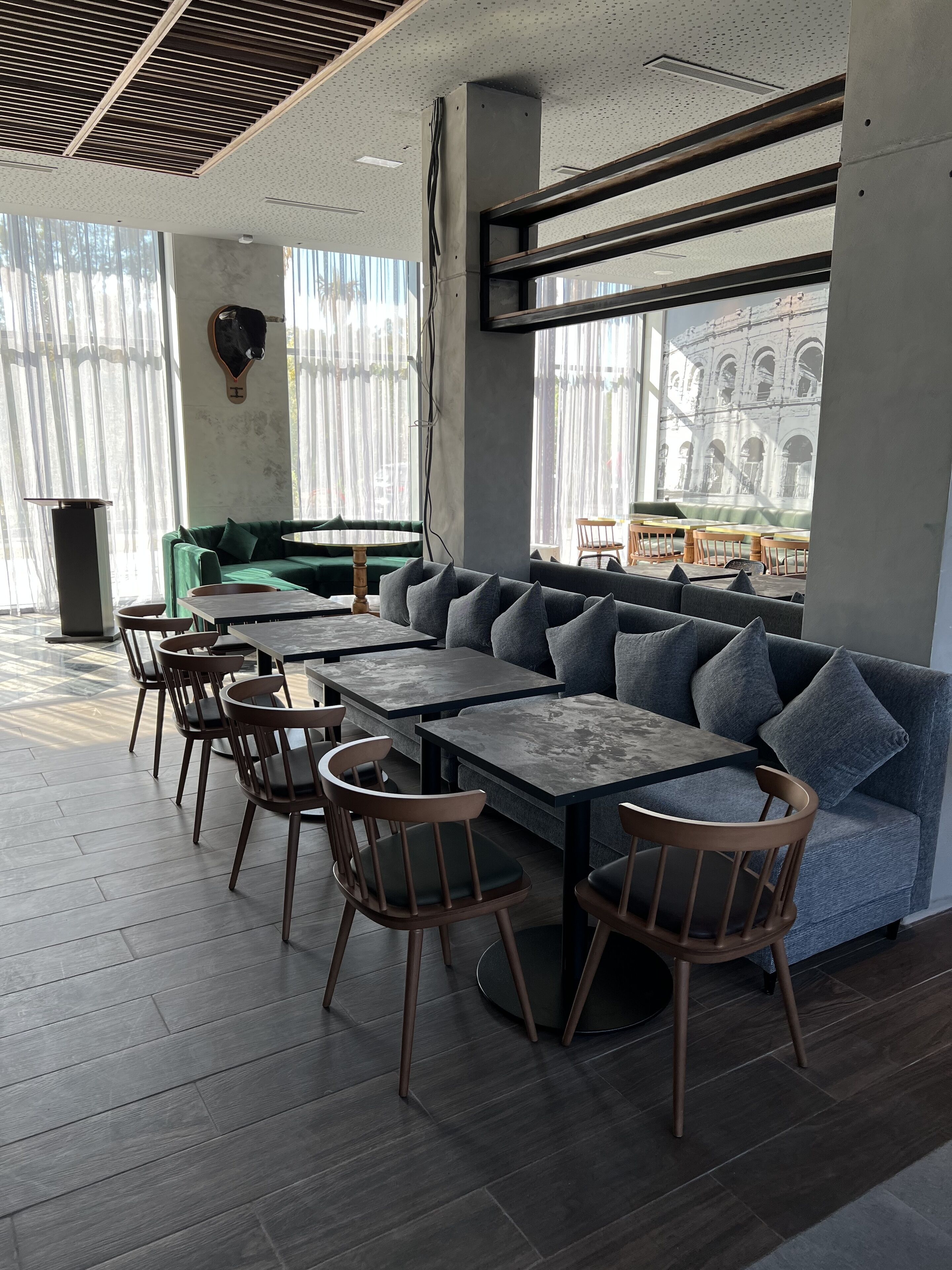 A modern interior with a long sofa, wooden chairs, and tables, large windows with a view of a building.