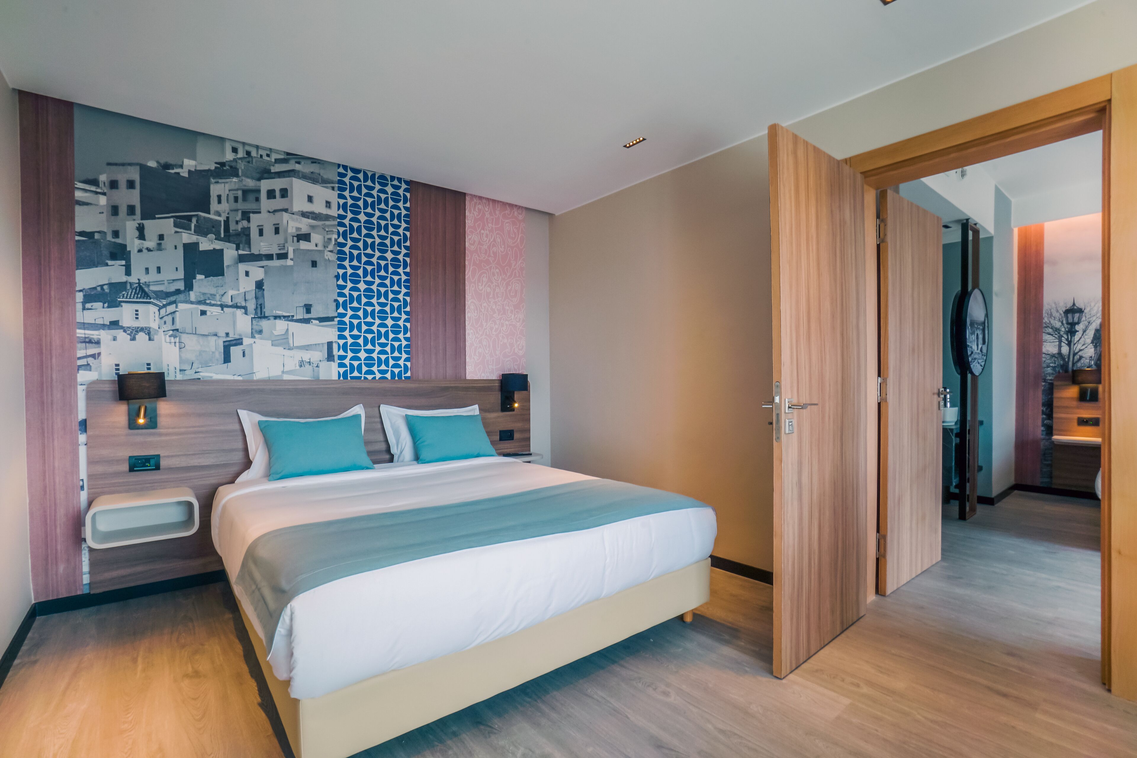A modern hotel room with a large bed, wooden headboard, and a cityscape mural on the wall.