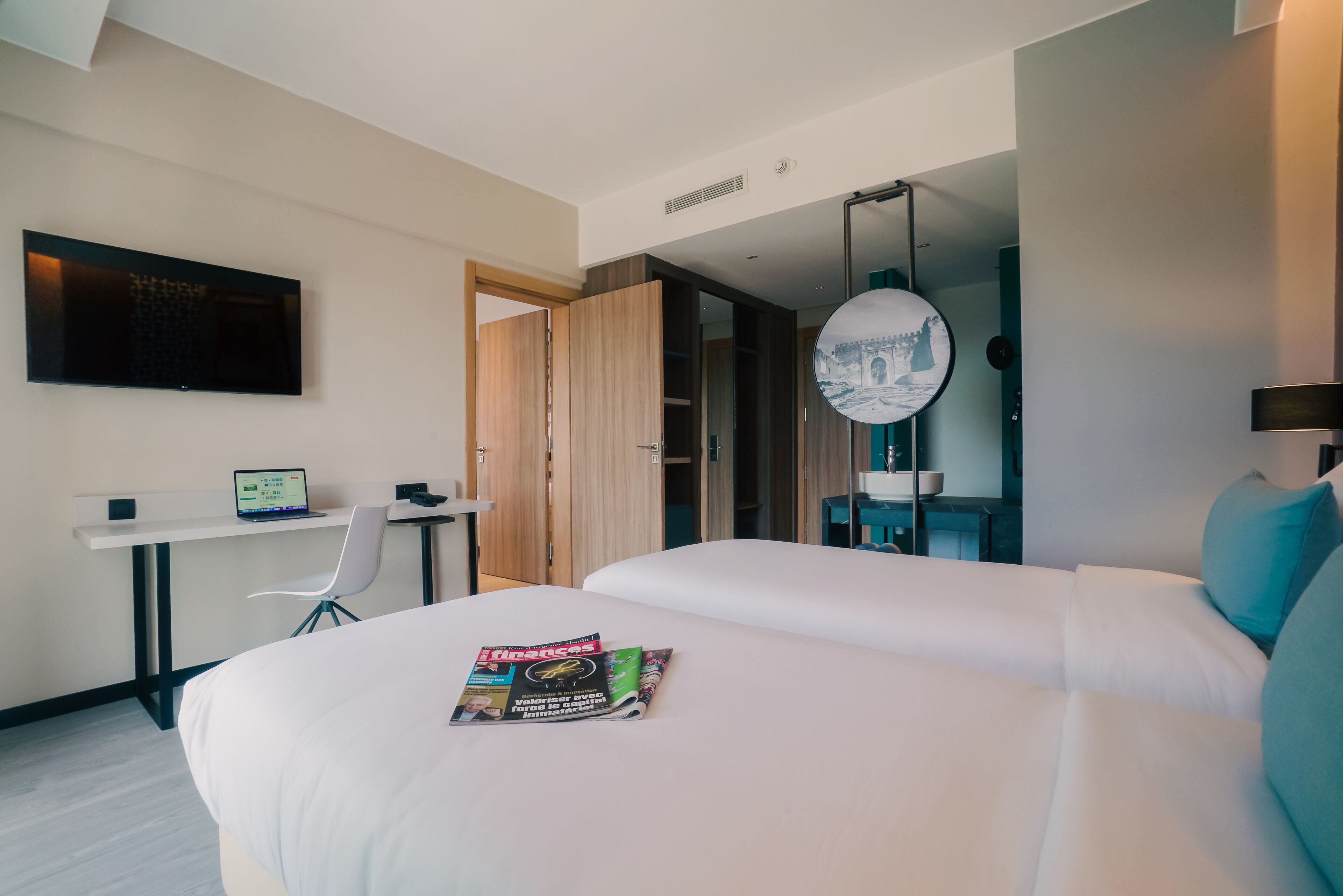 A modern hotel room with a large bed, a desk with a laptop, a TV mounted on the wall, and a circular mirror.
