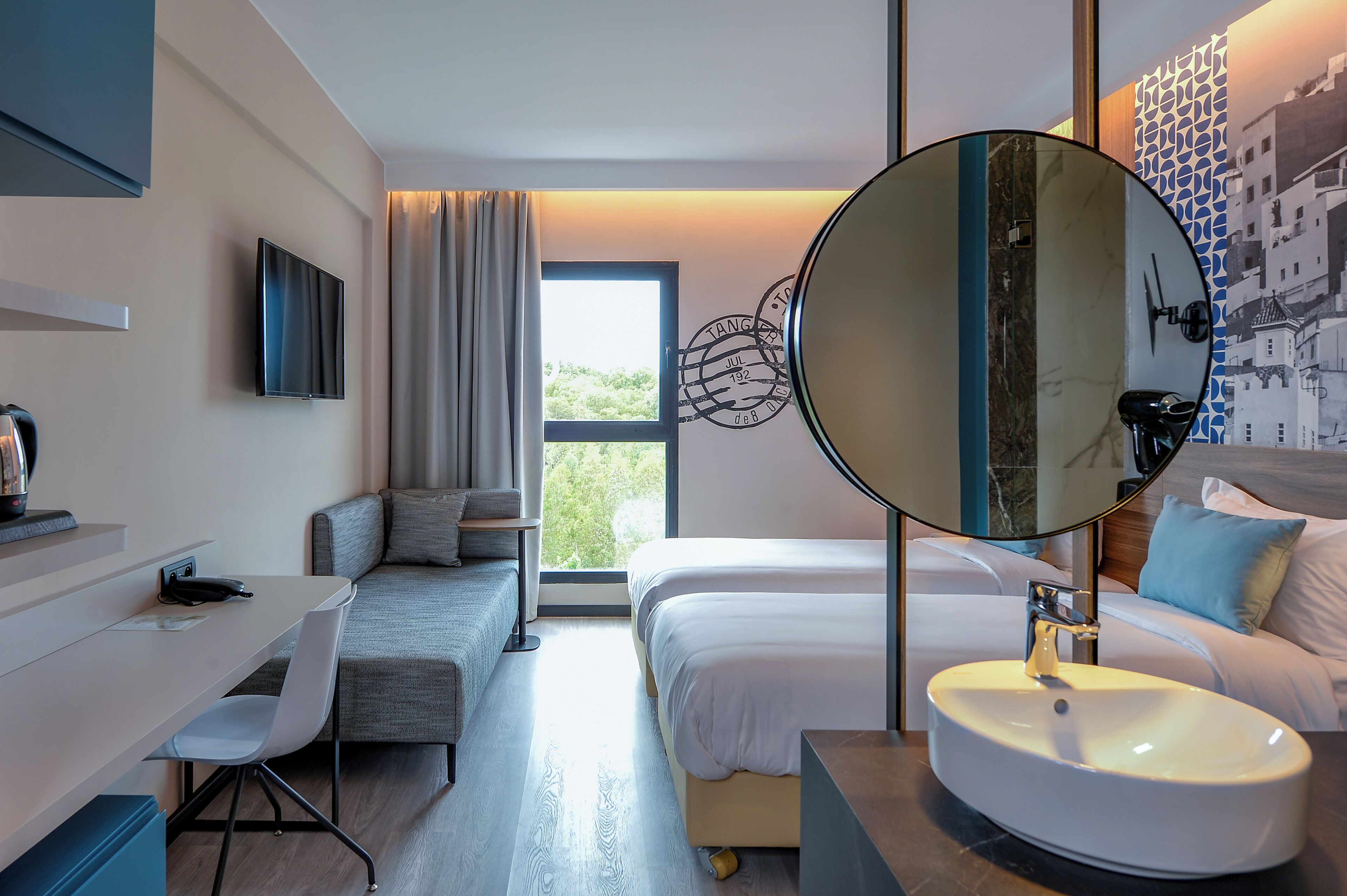 A modern hotel room with a bed, a desk, a chair, a large mirror, and a view of greenery through the window.