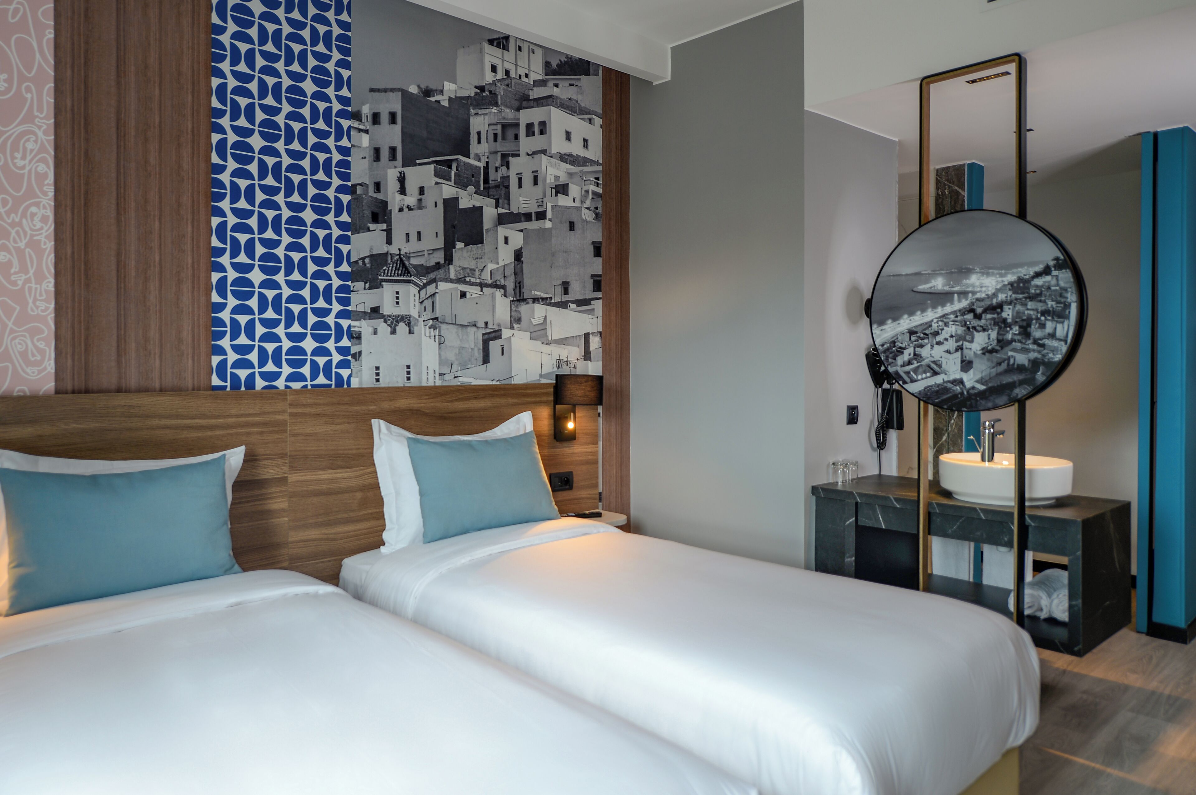 A hotel room with two beds, a large mirror, a round mirror, a sink, and a wall with a patterned design.