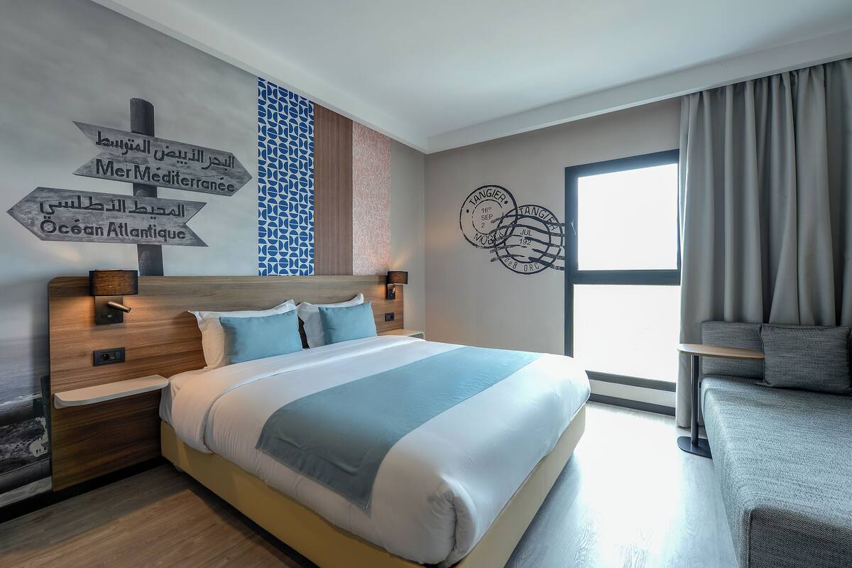 A modern hotel room with a large bed, a nightstand, a bedside table, a wall clock, and a window with curtains.