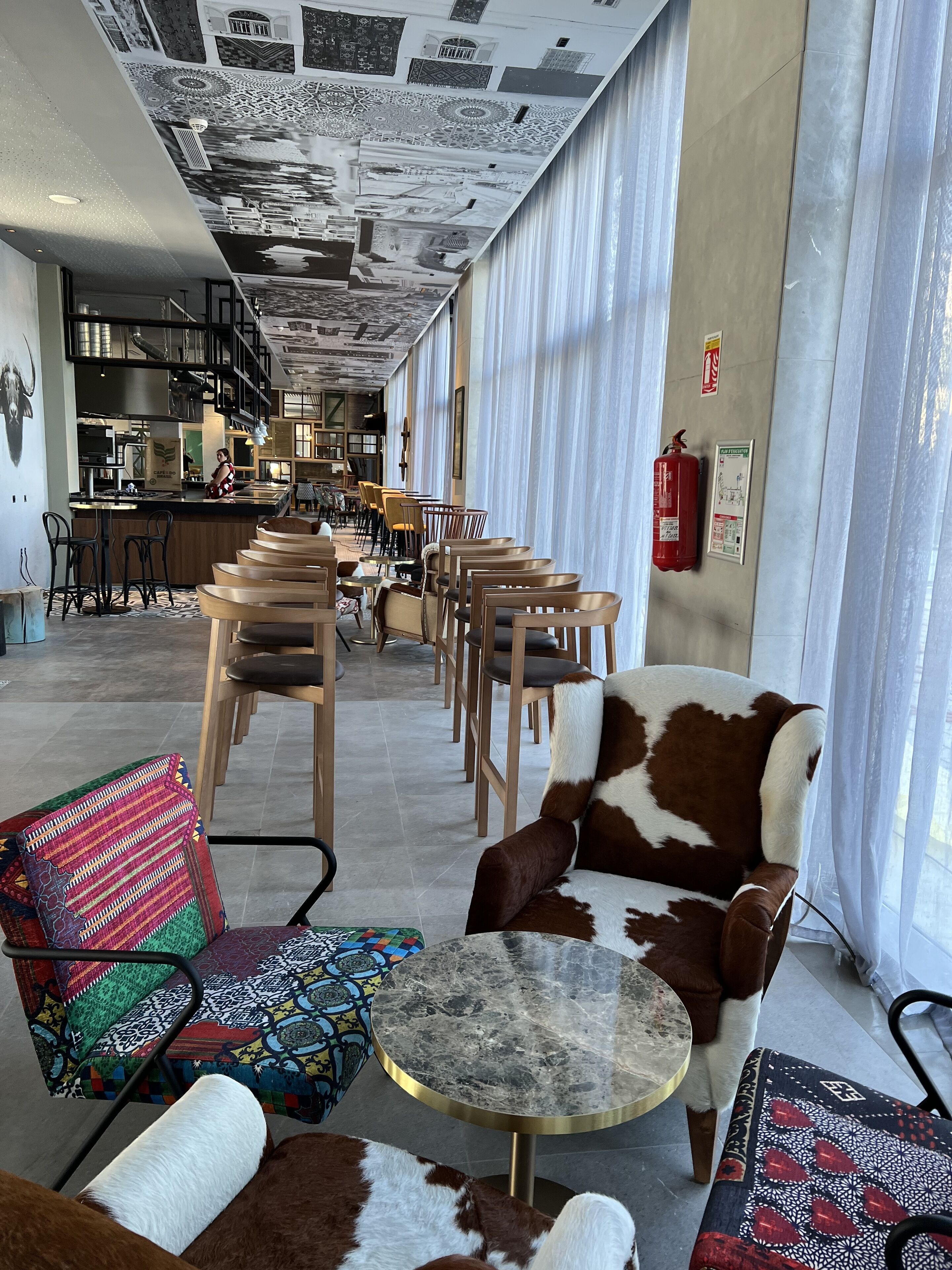 A modern interior with a long corridor leading to a bar area, featuring patterned ceiling, seating with patterned cushions, and a fire extinguisher on the wall.