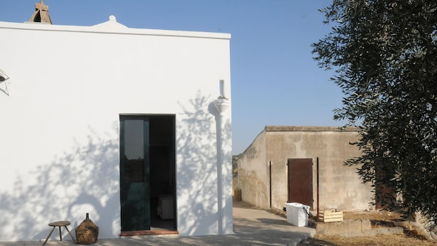 Un bâtiment blanc avec une porte, un petit tabouret et un panier à l’extérieur.