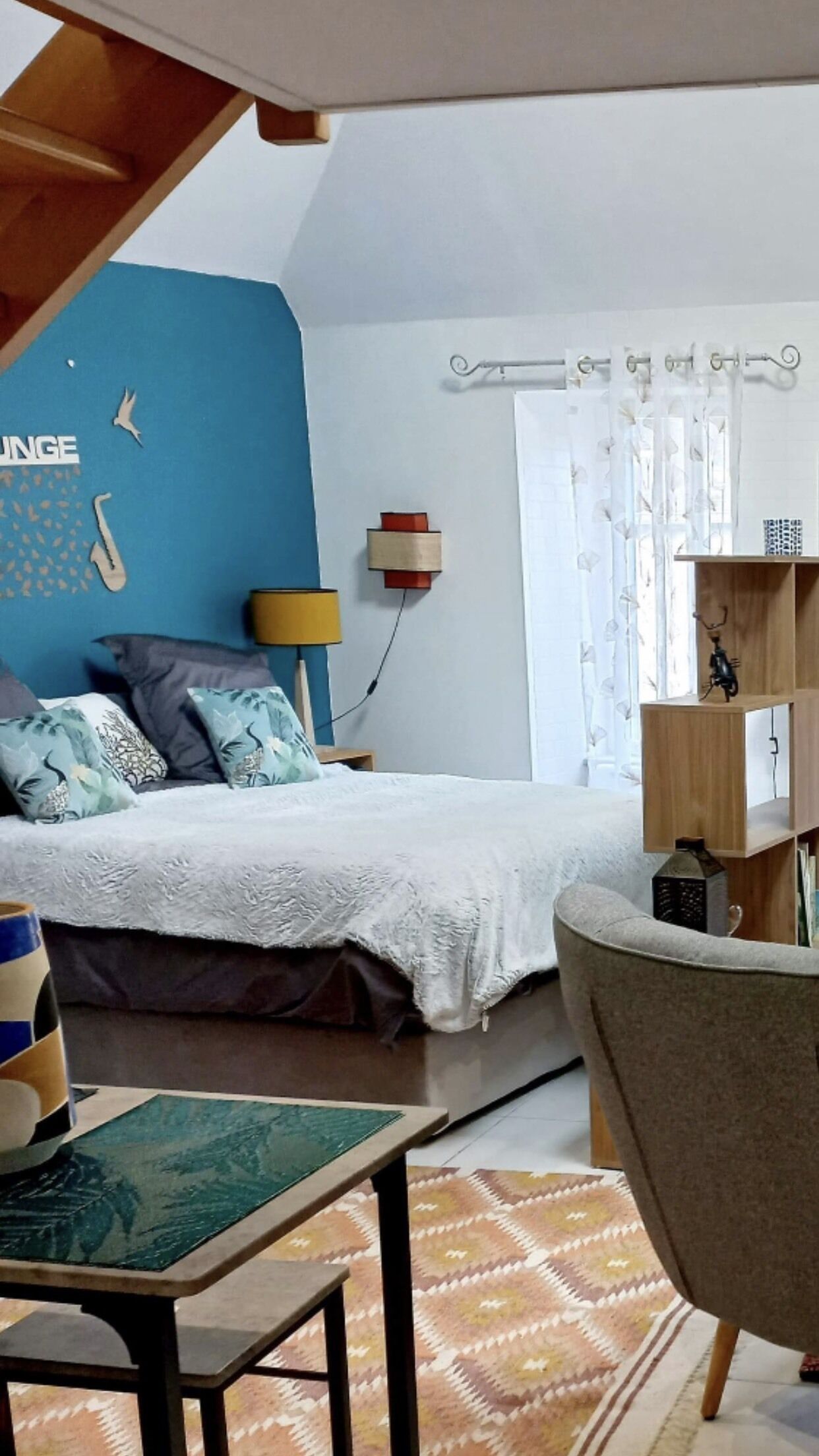 Une chambre à coucher avec un lit, une chaise, une table, une lampe, une étagère et une fenêtre avec des rideaux.