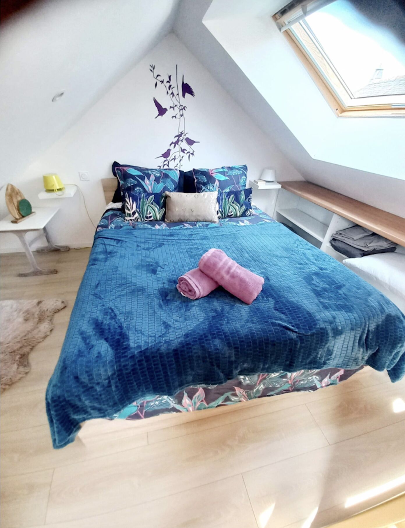 Une chambre avec une lucarne, un lit recouvert d’une courtepointe bleue, une table de chevet en bois et une petite table avec une lampe.