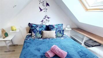 Un dormitorio con tragaluz, una cama con colcha azul, una mesita de noche de madera y una pequeña mesa con lámpara.