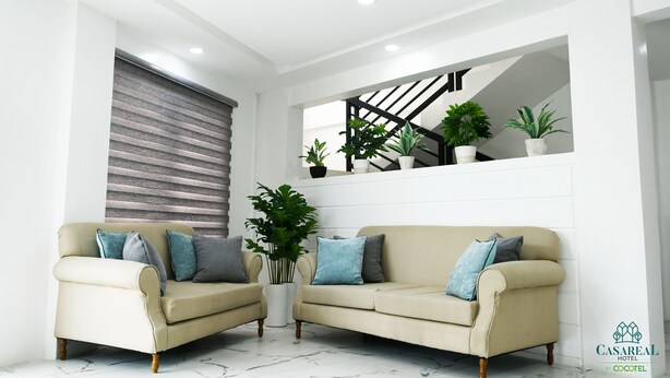 Een moderne woonkamer met twee beige banken, een groot raam met zonwering, een trap met potplanten en een witte muur met inbouwspots.