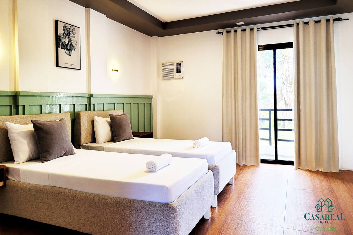 Ein Hotelzimmer mit zwei Betten, einem grünen Kopfteil, einem gerahmten Kunstwerk und einem Fenster mit Vorhängen.