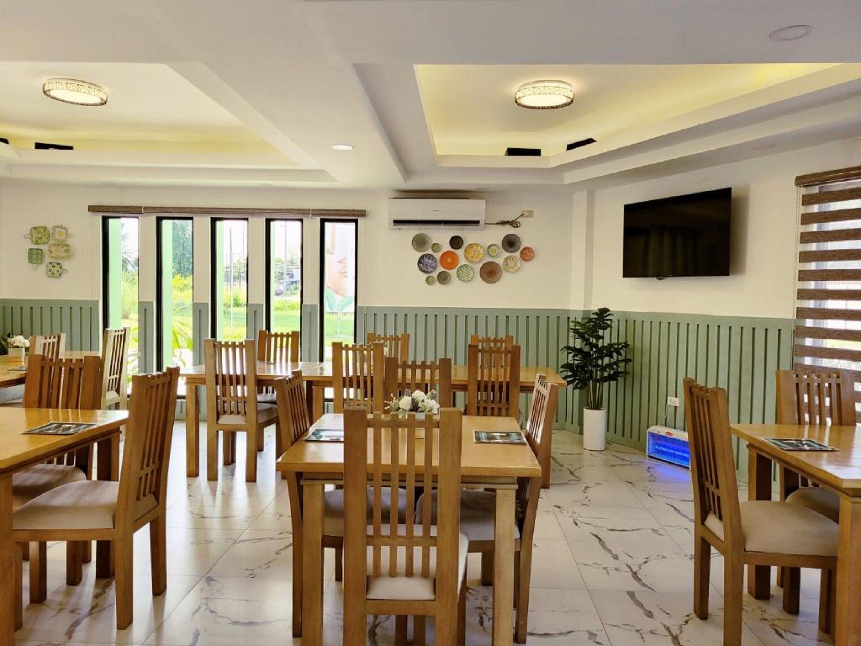 A dining area with wooden tables and chairs, a TV mounted on the wall, and a view of greenery outside.