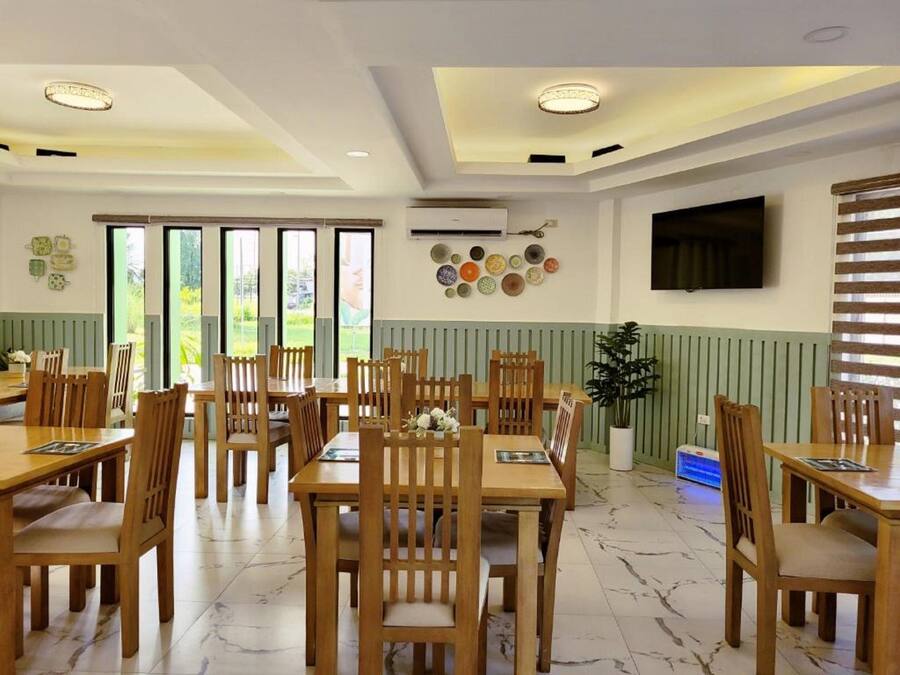 A dining area with wooden tables and chairs, a TV mounted on the wall, and a view of greenery outside.