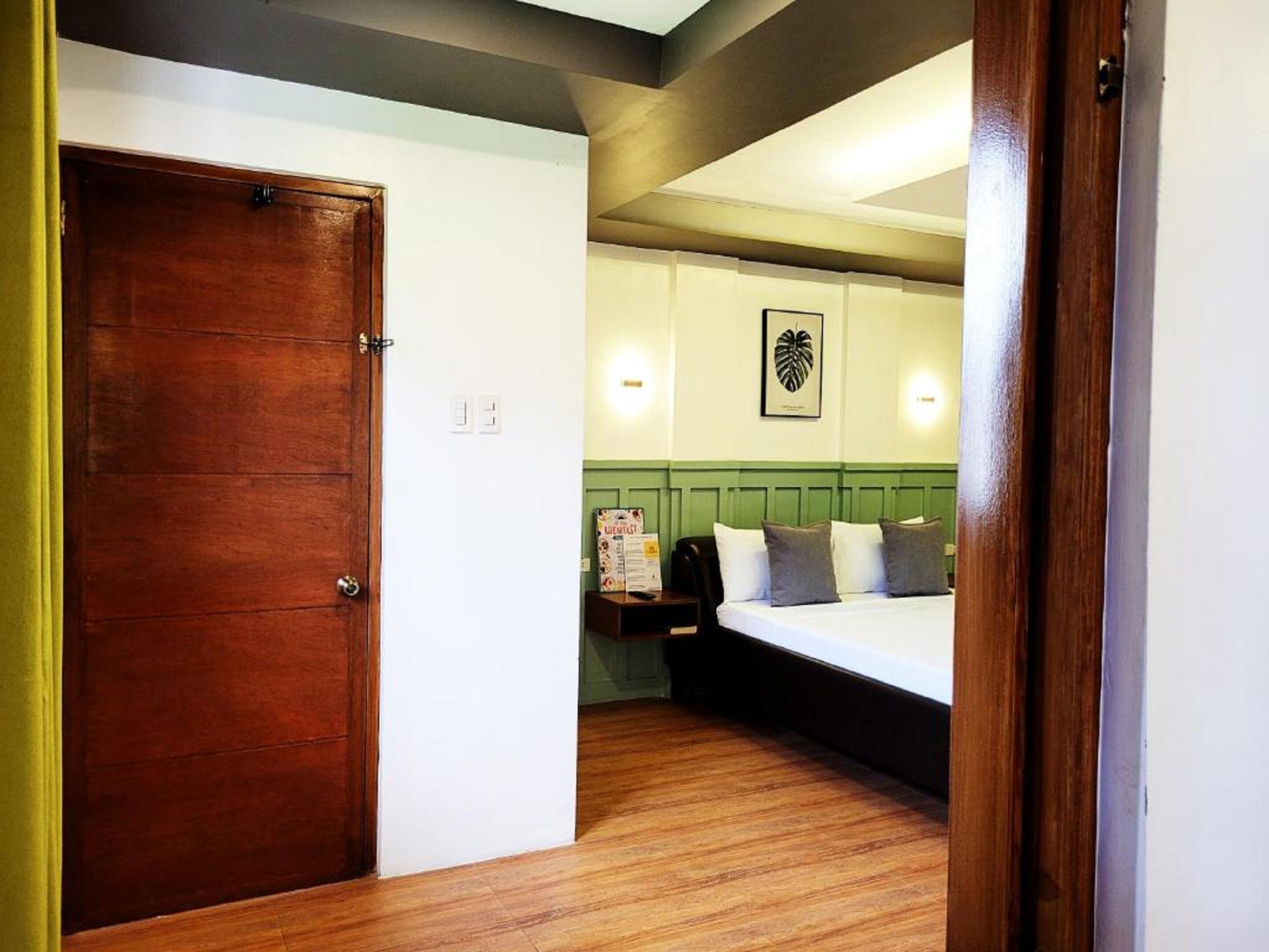 A hotel room with a wooden door, a bed with pillows, a green headboard, and a wall-mounted light fixture.