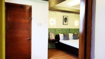 A hotel room with a wooden door, a bed with pillows, a green headboard, and a wall-mounted light fixture.