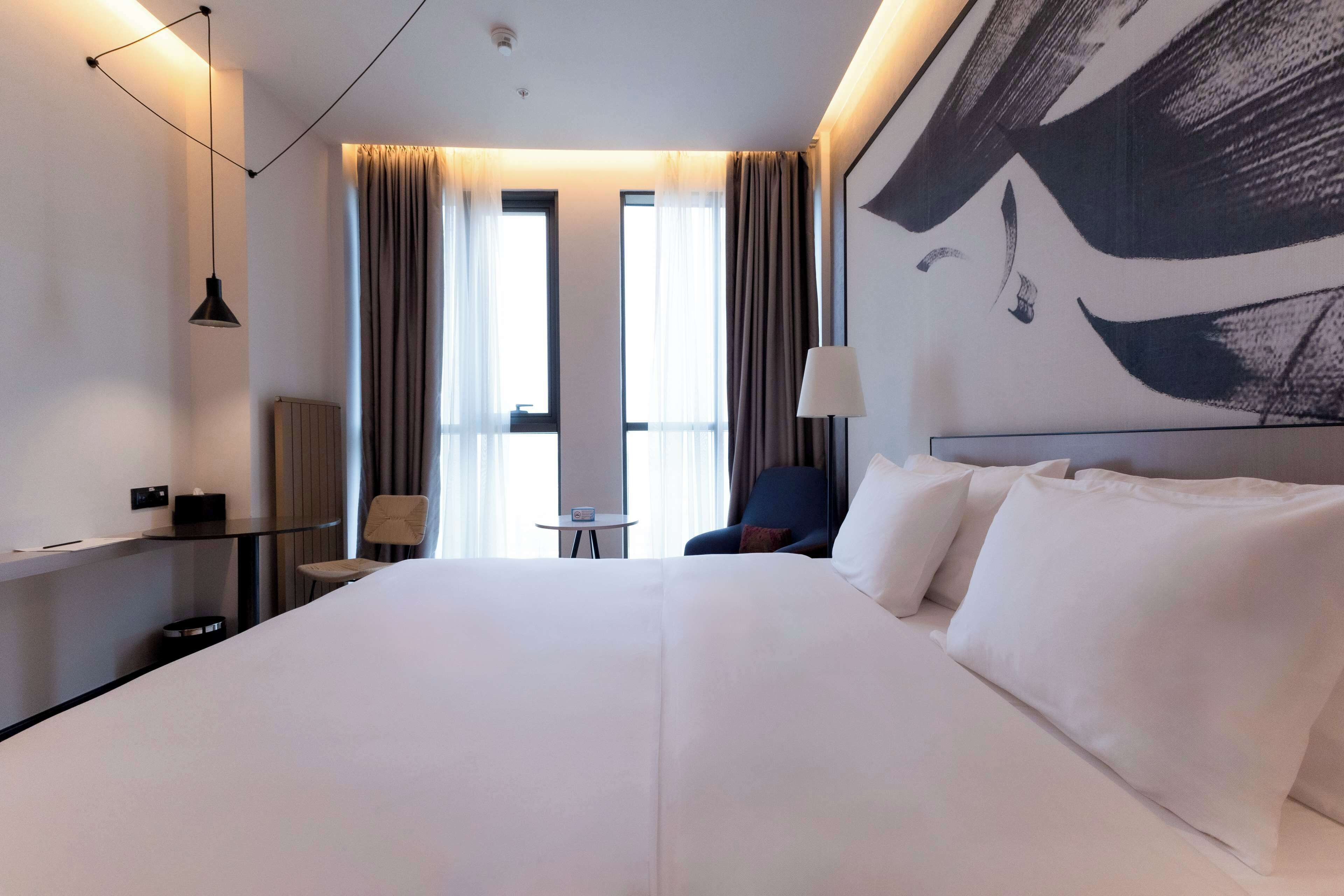 A hotel room with a large bed, a desk with a chair, a small table, and a large window with curtains.