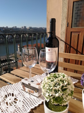 A balcony with a view of a river and buildings, a bottle of wine, two glasses, a box of chocolates, and a potted plant.