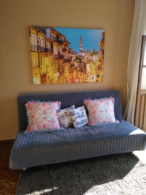 A room with a blue sofa bed, two patterned pillows, a large cityscape painting on the wall, and a window with curtains.