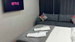 A hotel room with a bed, a sofa, a TV displaying the Netflix logo, and a ceiling with recessed lighting.