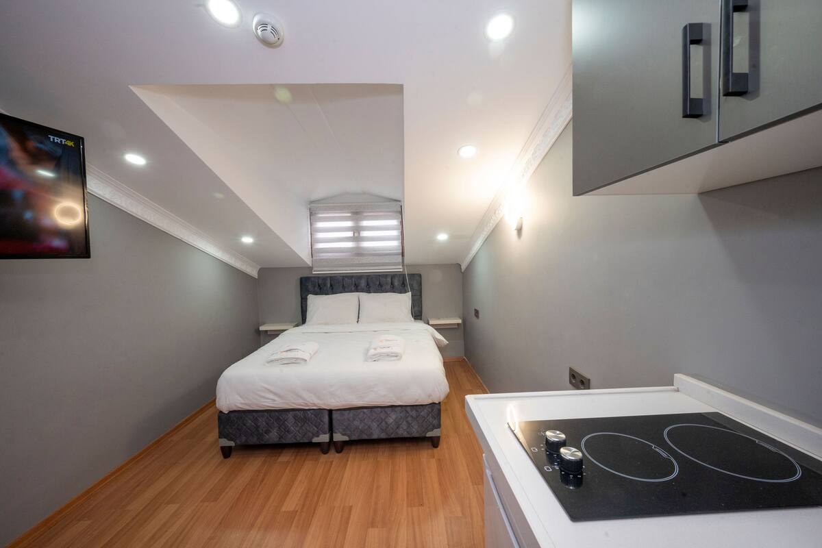 A modern hotel room with a bed, a built-in stove, and a television on the wall.