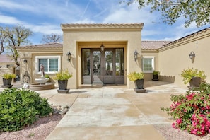 A house with a beige facade, a tiled roof, and a front entrance with a decorative door. There are potted plants and a fountain in the front yard.