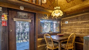 Interior de una cabaña de madera con una mesa de comedor y sillas, una ventana que ofrece vistas a árboles y un extintor en la pared.