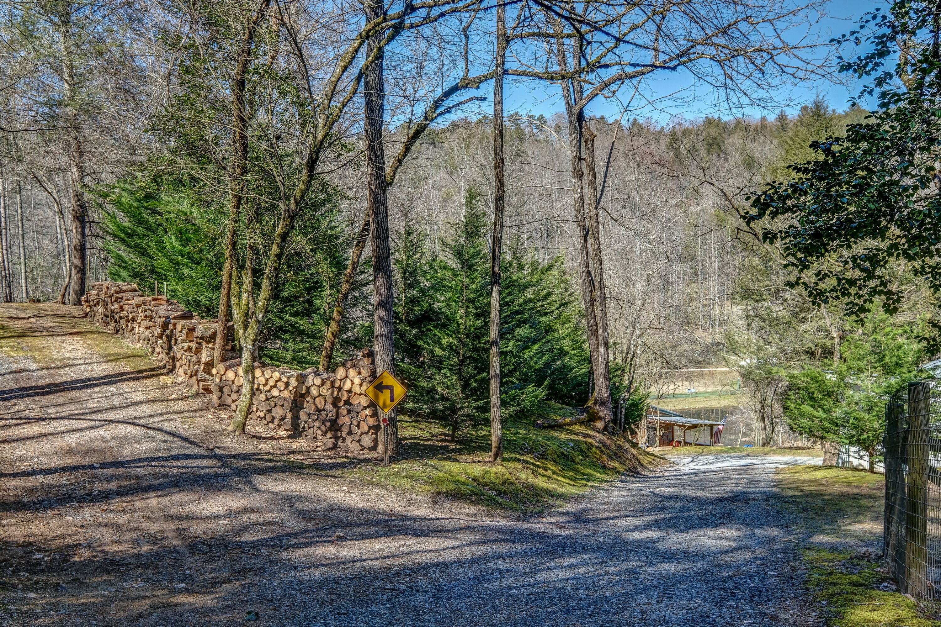 A gravel road winding through a forested area with a stone wall and a yellow warning sign.