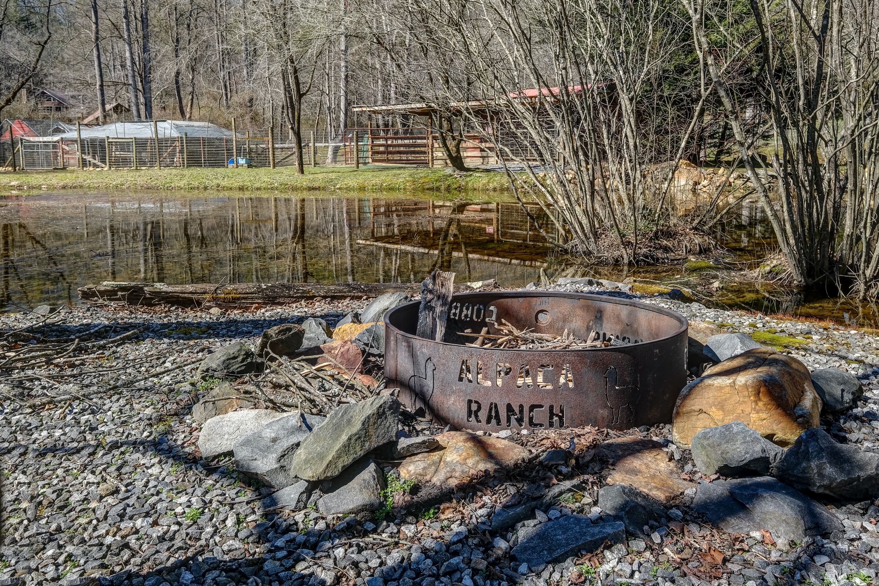 A rustic outdoor fire pit labeled "ALPACA RANCH" surrounded by rocks and logs, with a pond and trees in the background.