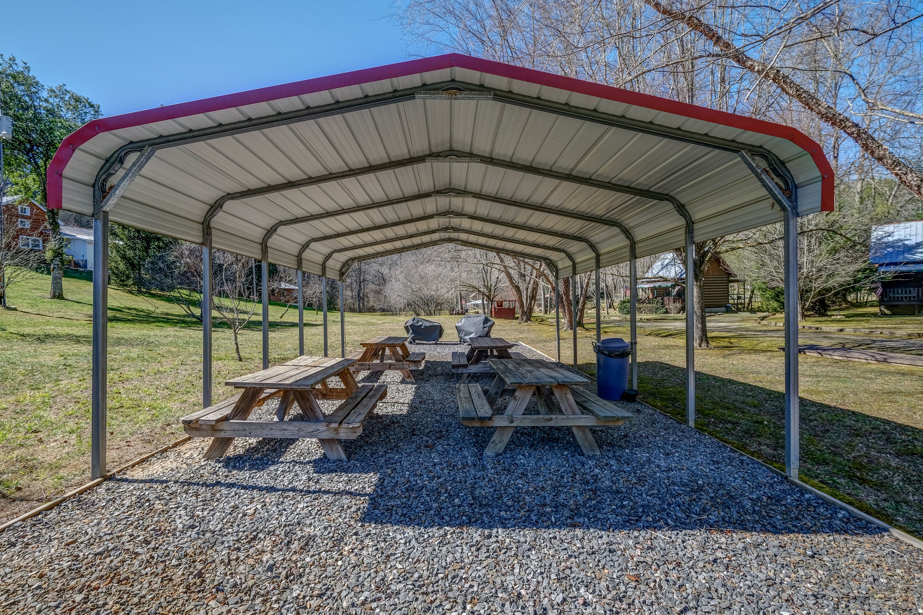 A covered picnic area with benches and picnic tables, surrounded by gravel and trees.