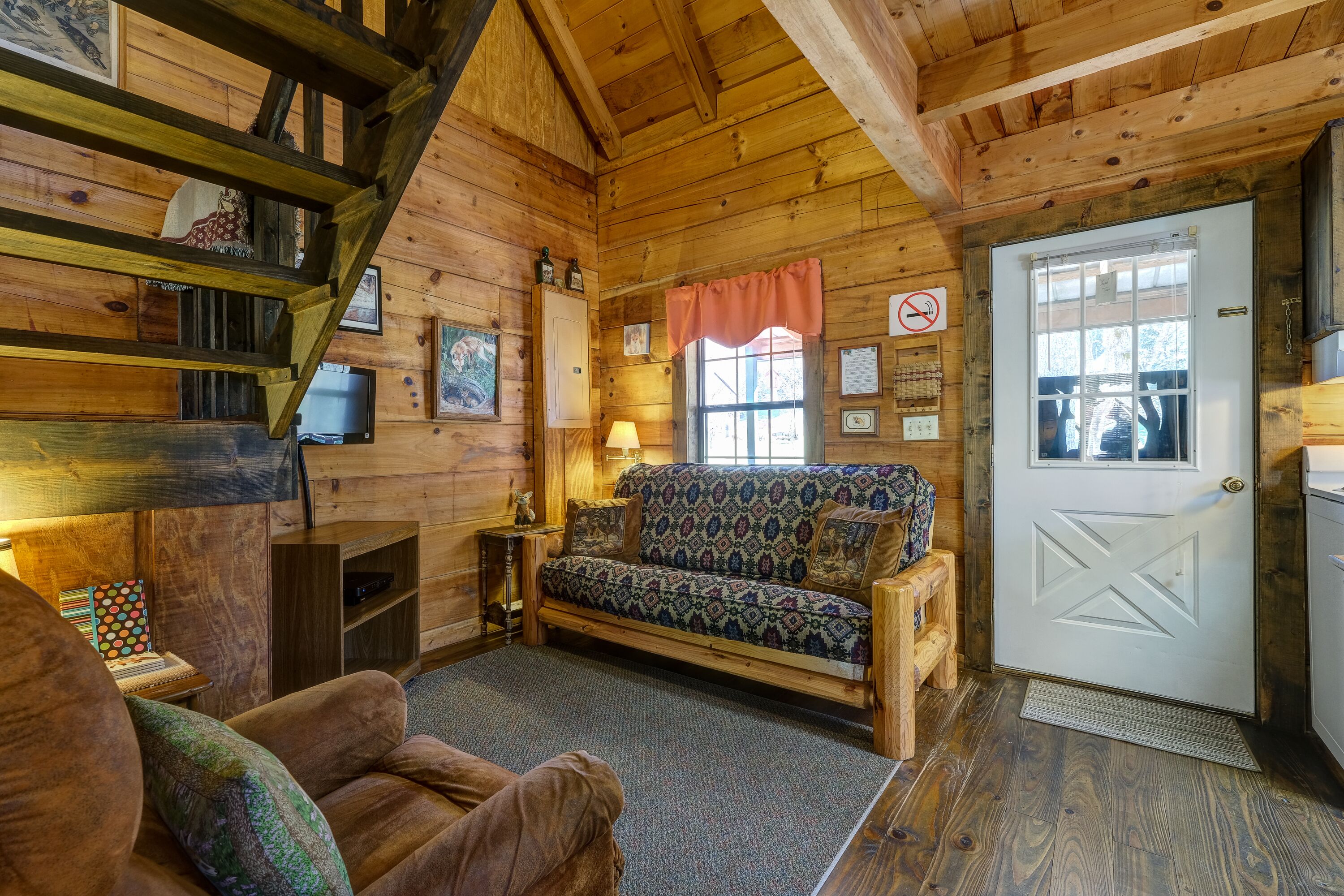 A cozy log cabin interior with a sofa, a wooden staircase, a fireplace, and a door with a window.