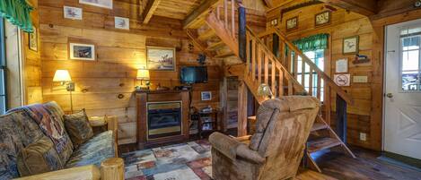 A cozy log cabin interior with a wooden staircase, a fireplace, a television, and a plush armchair.