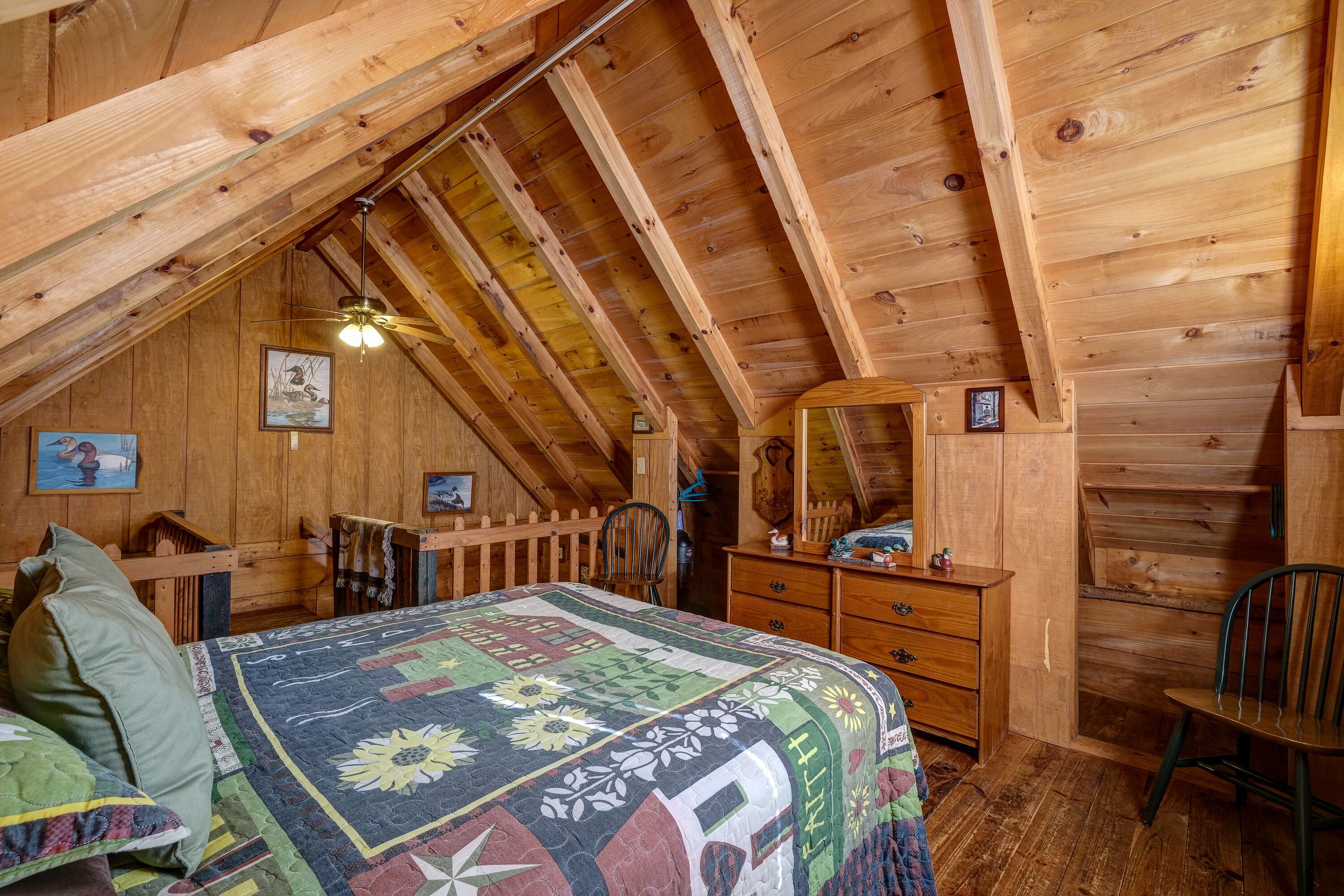 Un dormitorio acogedor en el ático con techo de madera, una cama con colcha colorida, un aparador, una silla y un espejo.