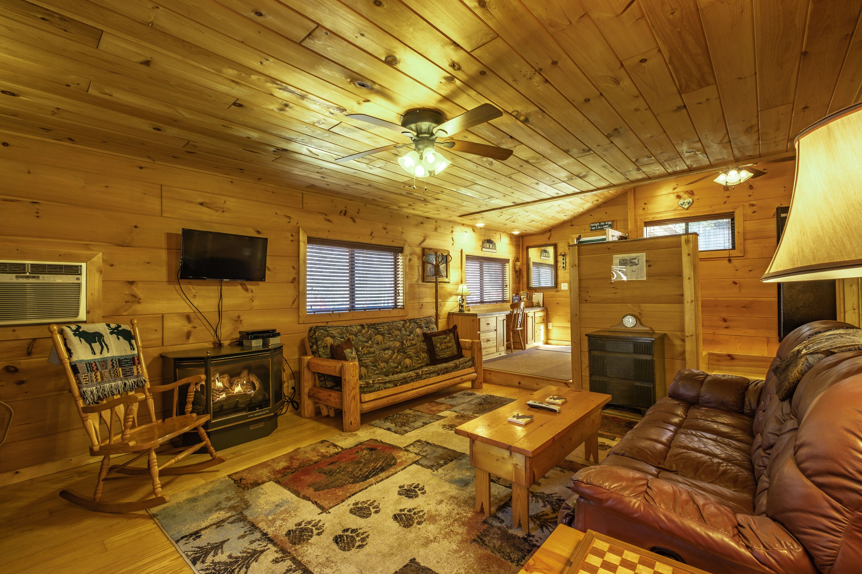 A cozy cabin interior with a fireplace, wooden furniture, a television, and a ceiling fan.