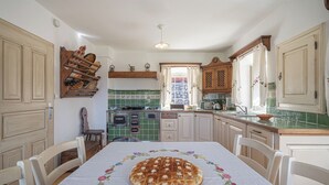 A kitchen with a table set for dining, a hanging rack with dishes, and a window with curtains. - Traditional House Kal Garden & Jacuzzi (Semič)