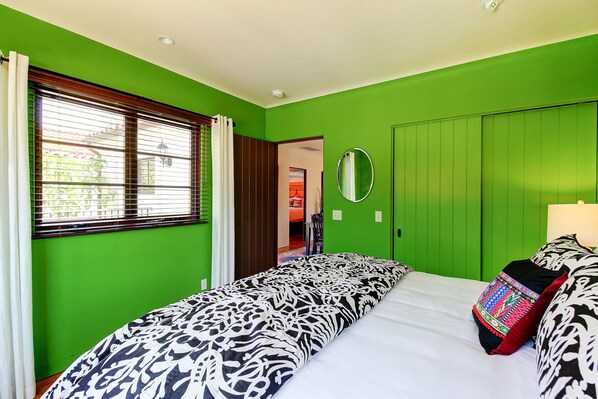 A bedroom with a green wall, a bed with patterned bedding, a bedside mirror, and a window with blinds. - West Beach Villas 5 & 6 - 2 villa rental (entire property) - 2 blocks to beach, State St! (Santa Barbara)